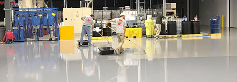 Workers applying epoxy floor coating in a large industrial space with equipment in the background.
